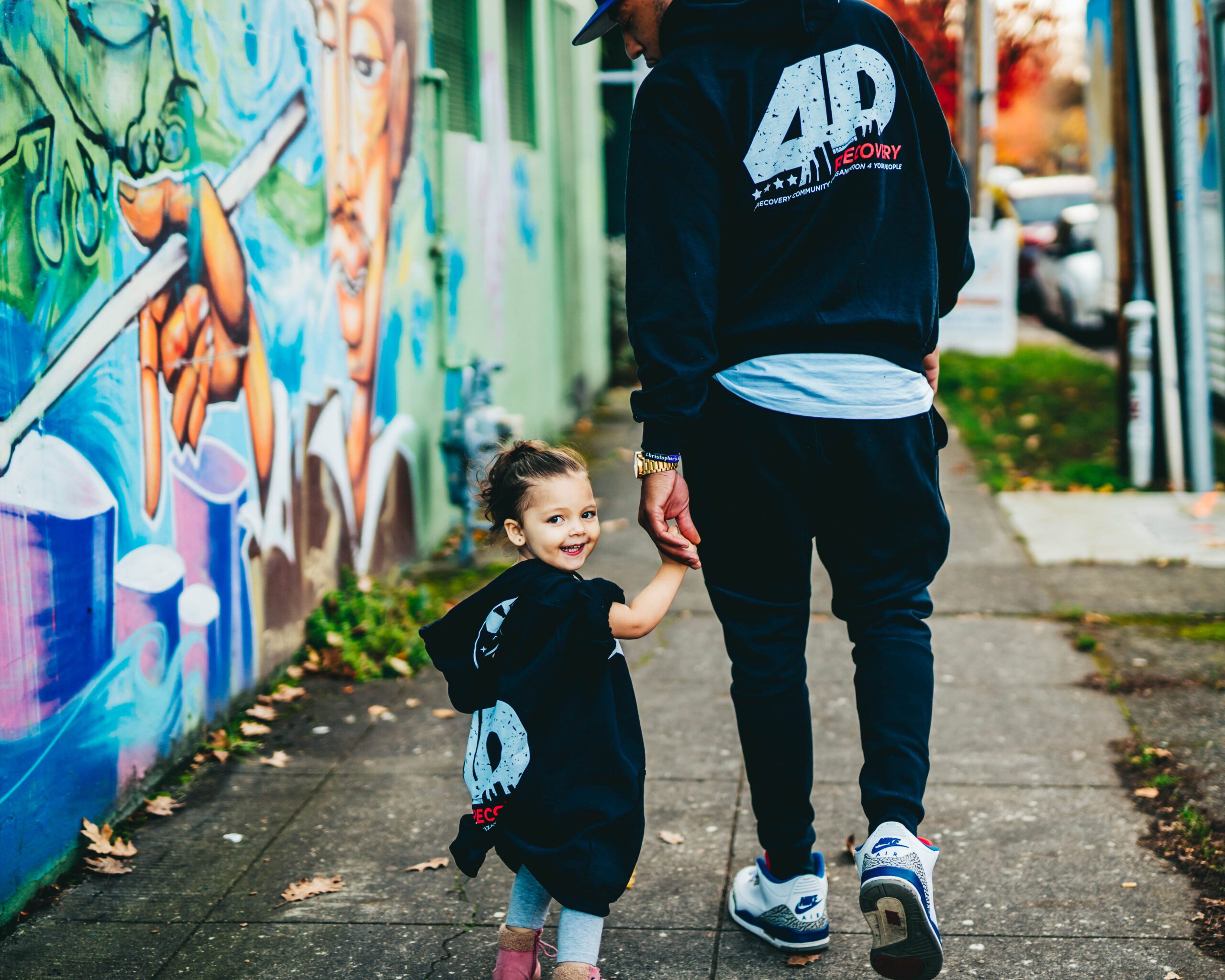 Young man walks with child holding hands next to graffiti wall, both wear 4D sweatshirts.